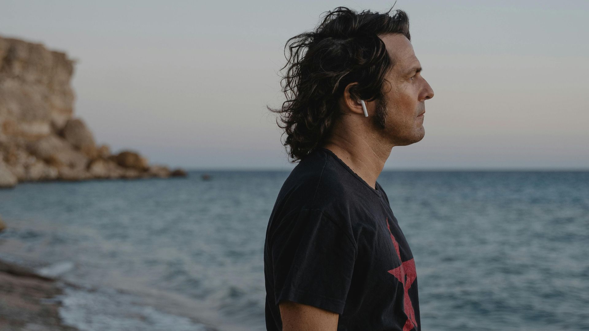 A man in profile wearing earbuds gazes at the ocean during sunset, exuding peace.