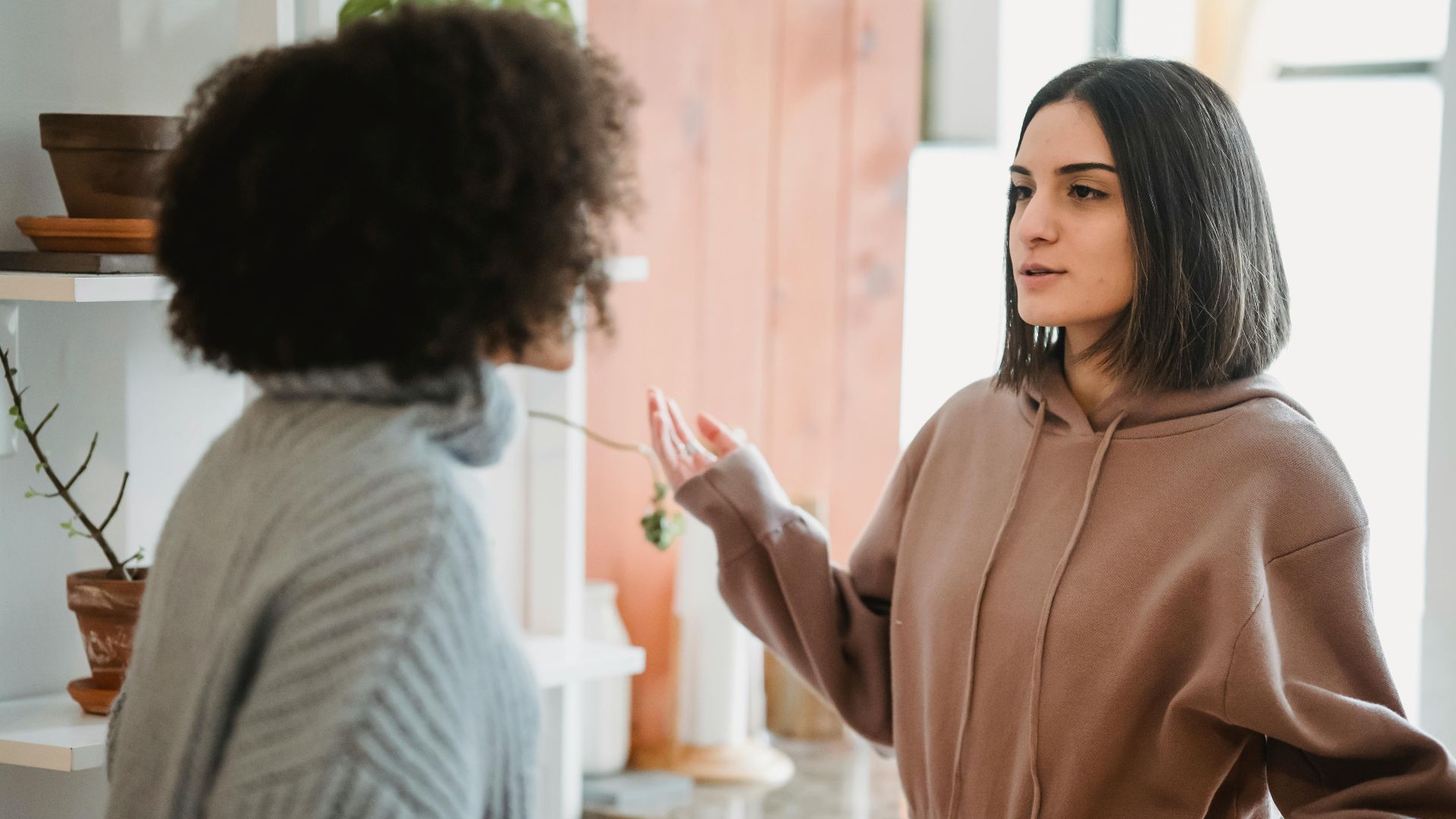 Serious woman in casual wear having argument with anonymous female with curly hair while standing in light room at home