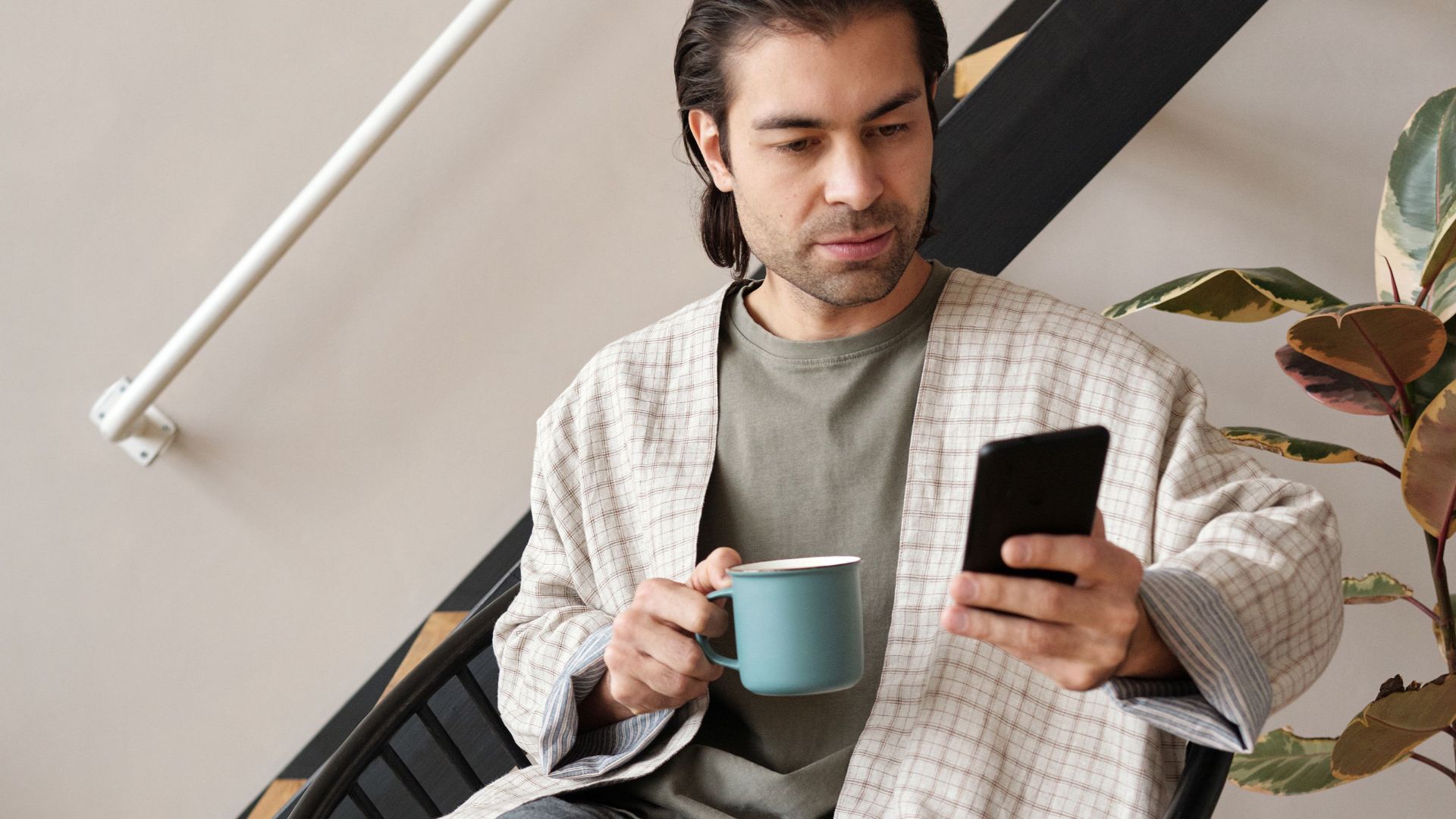 Man seated indoors, holding a coffee mug and smartphone, relaxing in comfortable attire.