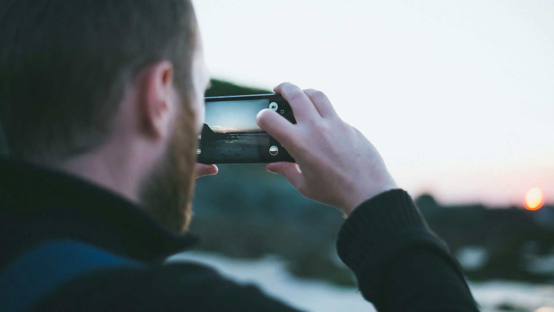 A man with a beard photographing a serene beach sunset using a smartphone.