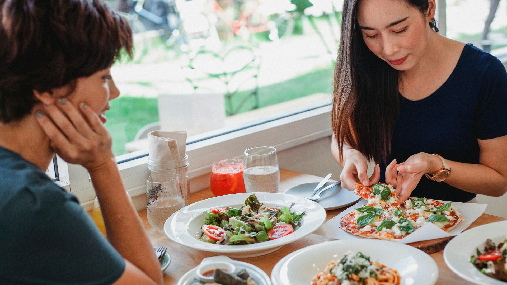 Content Asian woman taking slice of pizza while sitting at table near friend with delicious salad and pasta in restaurant