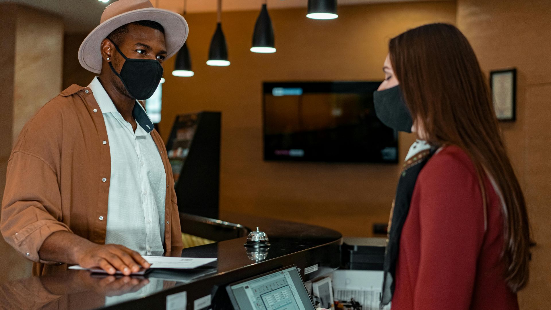 A man and woman in face masks checking in at a hotel reception, emphasizing safety protocols.