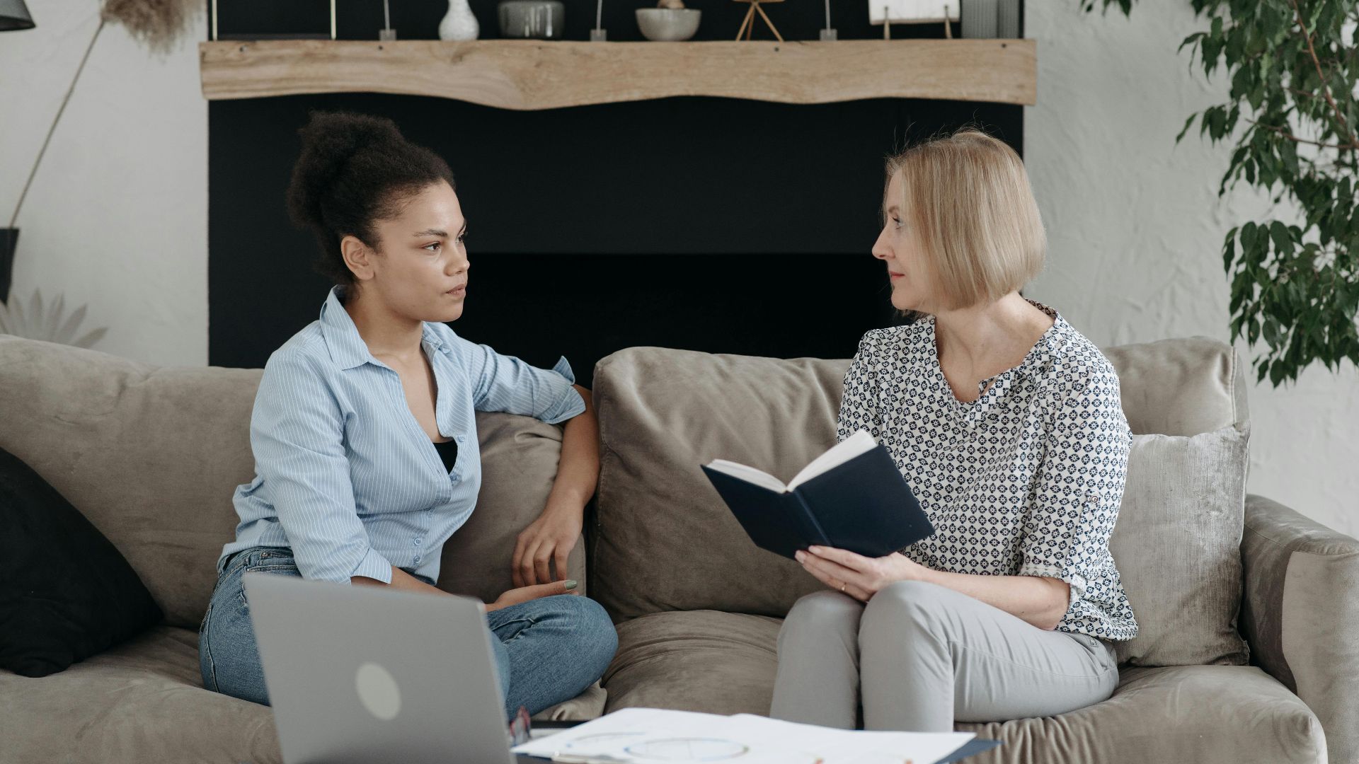 Two women sitting on a sofa having a thoughtful discussion, one holding a book.