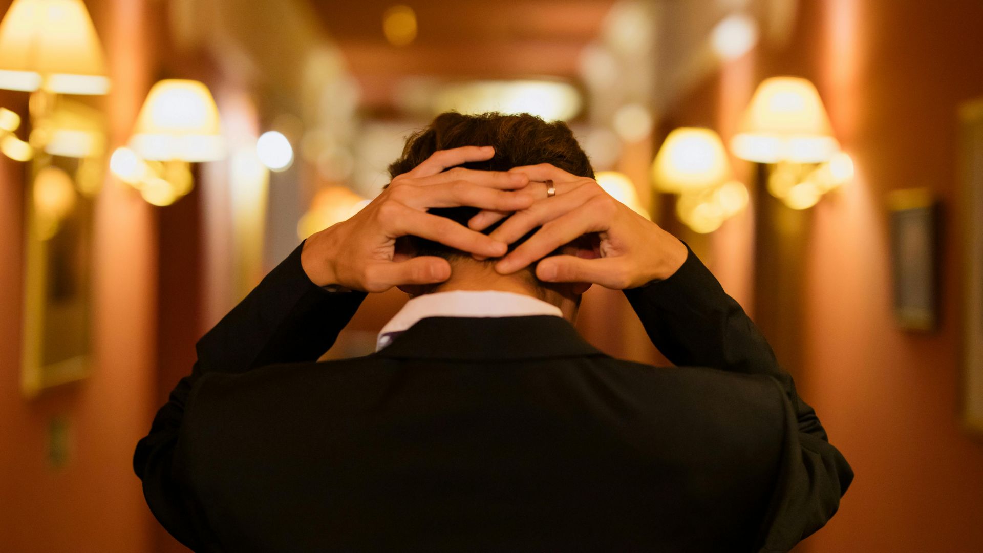 Back view of a man in a suit standing in a warmly lit hotel corridor.