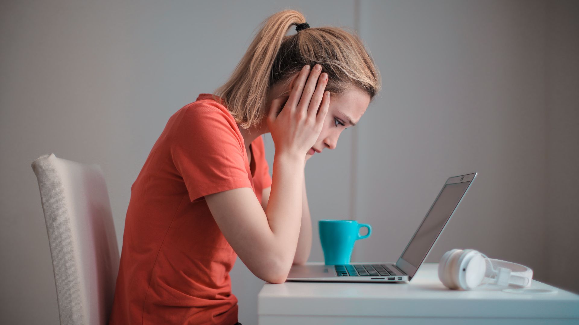 Side view of thoughtful worried female student in casual clothes focusing on screen and trying to solve problem while sitting at table with blue mug of hot drink and headphones and working on laptop at home