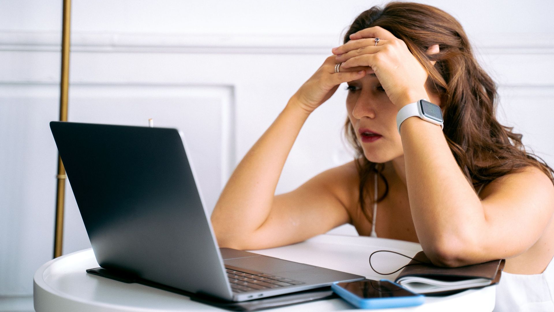 Woman feeling stressed while working on a laptop, using smartphone and smartwatch at a desk.