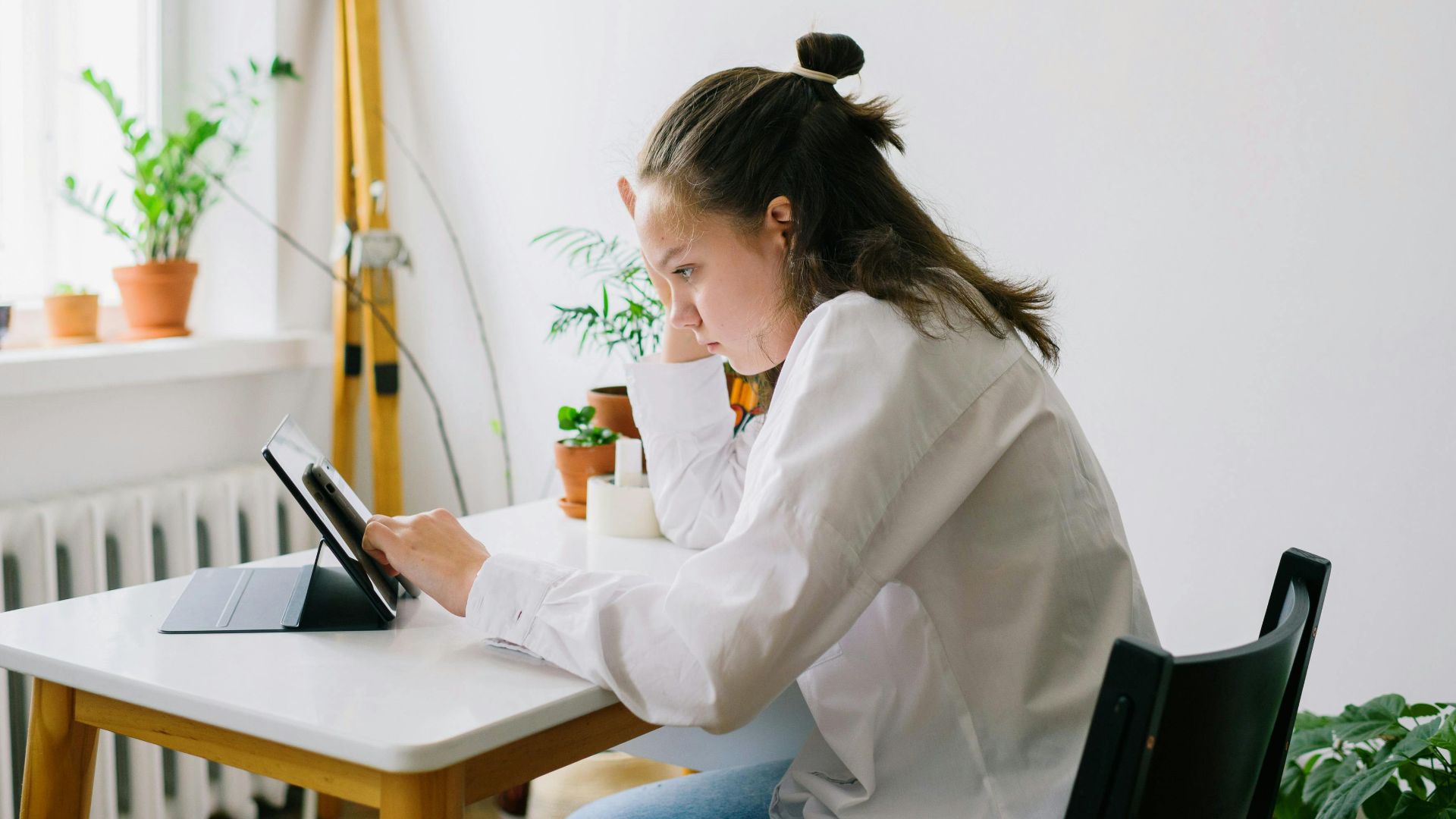 Young woman in casual white shirt working on tablet at home desk surrounded by plants.
