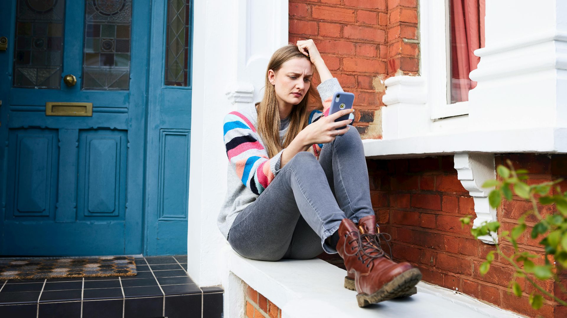 Young woman using phone outside red brick house with blue door, looking thoughtful.