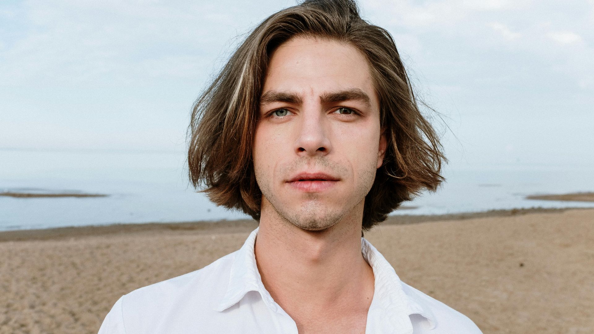 A young man poses on a sandy beach under a clear blue sky.