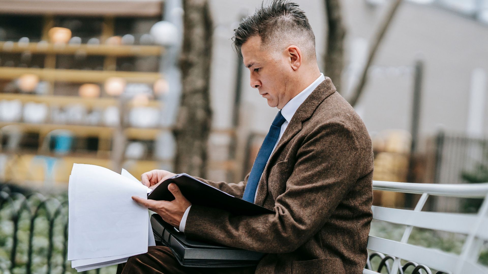 A professional man sitting on a park bench reviewing documents. Business attire, focused and engaged.