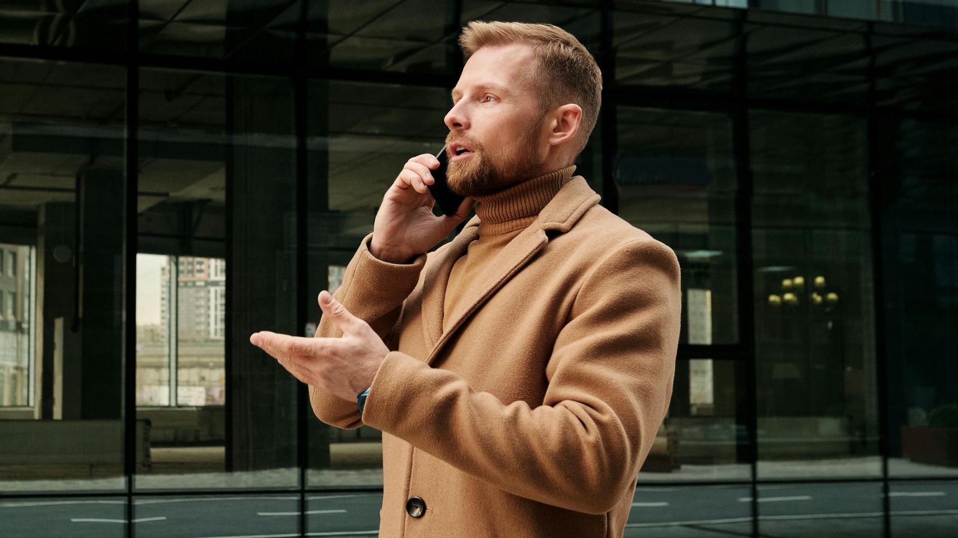 Confident businessman in a brown coat talks on phone outdoors in city.