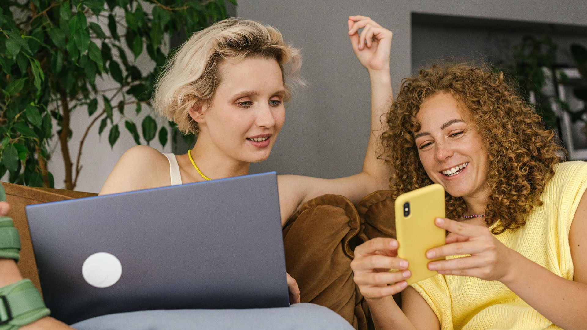 Two women sitting on a couch, engaging with a smartphone and laptop in a relaxed indoor setting.