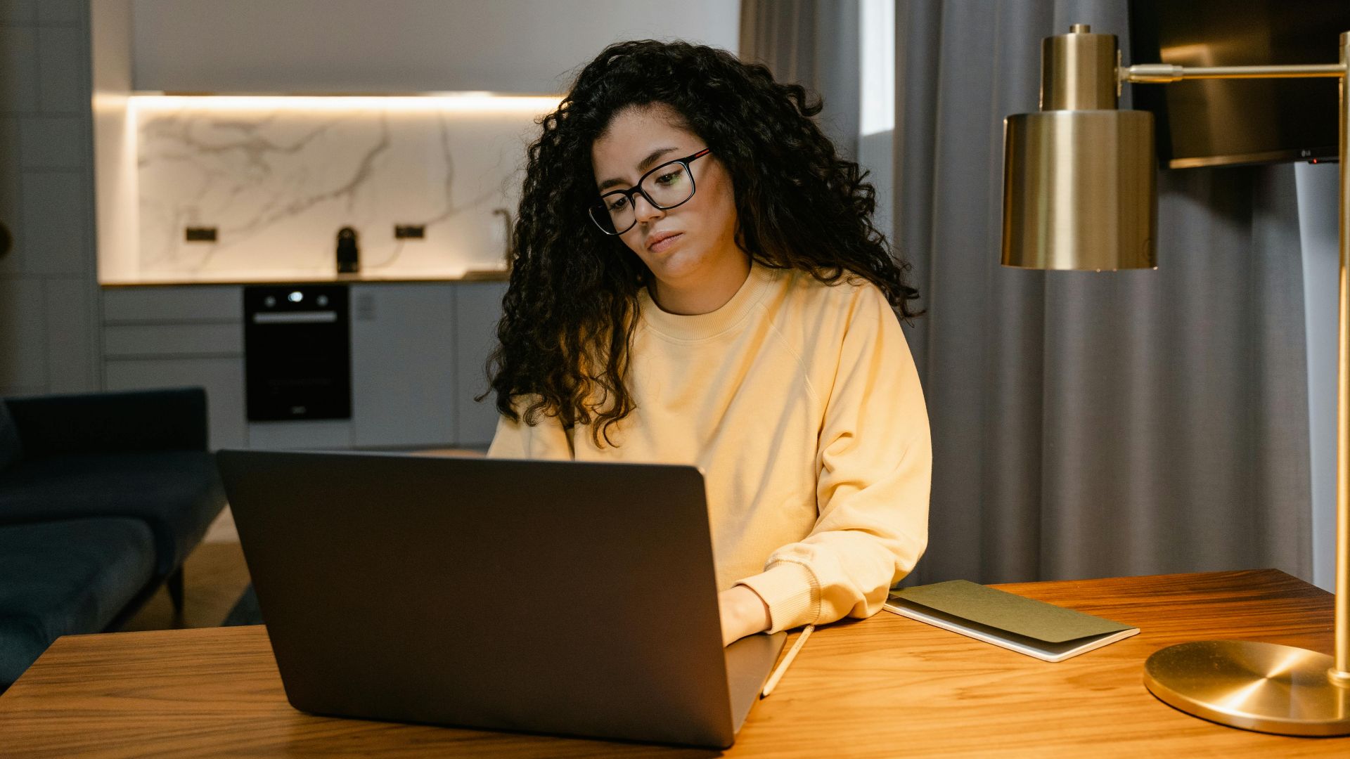 Woman in a yellow sweatshirt using a laptop at home, focusing on remote work in a cozy, modern interior.