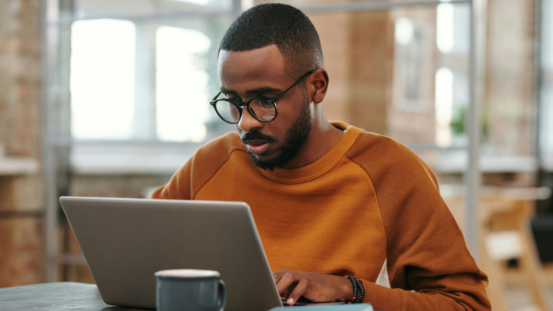 Young man with glasses works on laptop at a wooden table indoors. Warm, productive atmosphere.