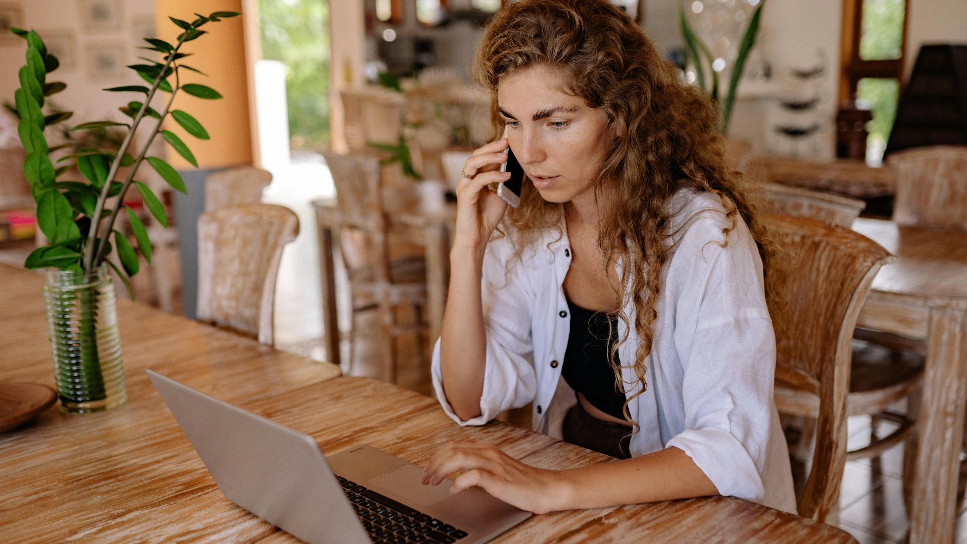 A woman in a bright room using a laptop and phone, blending work with home comfort.