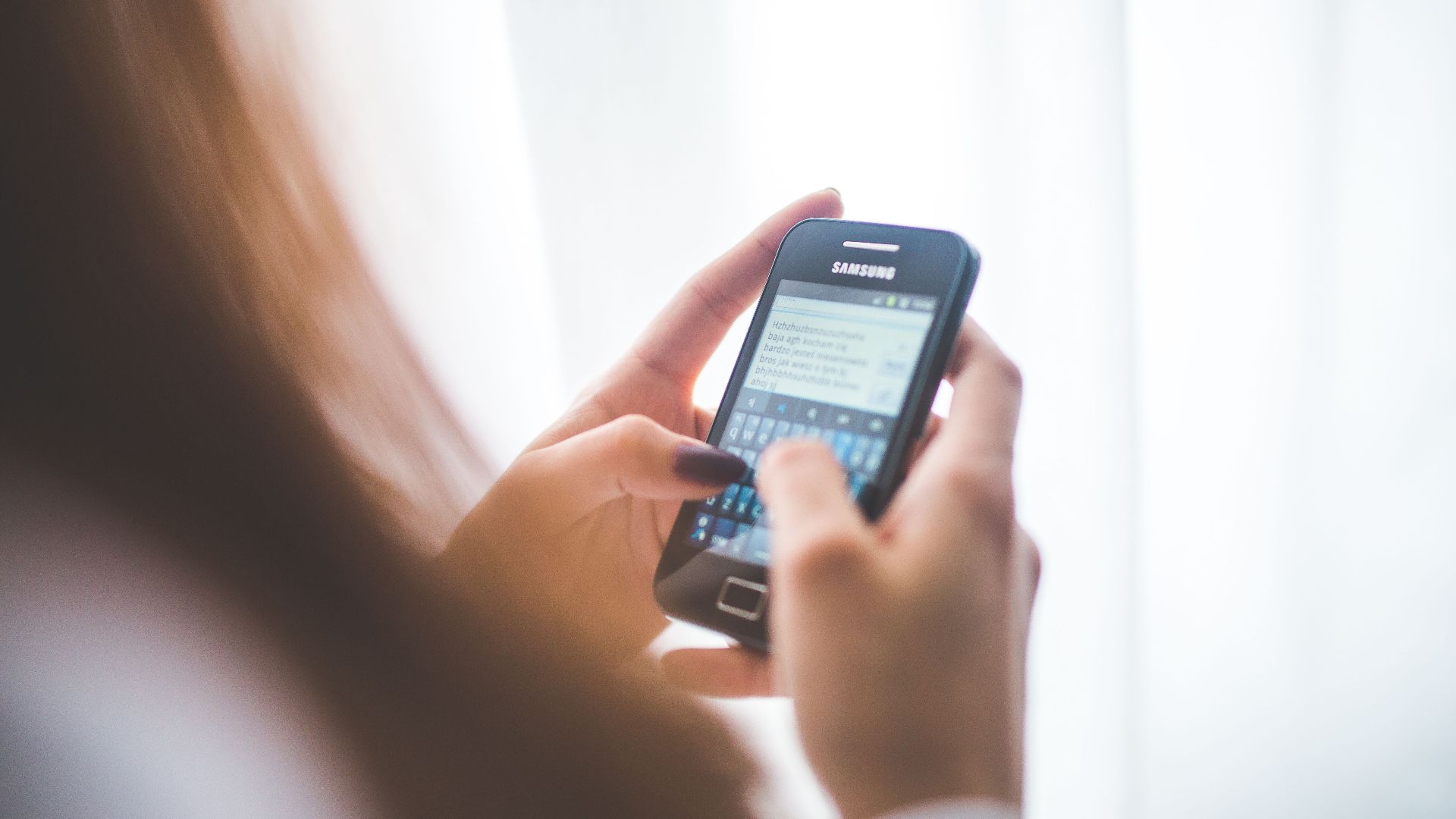 Close-up of a young woman typing on a Samsung smartphone at home. Bright indoor lighting.