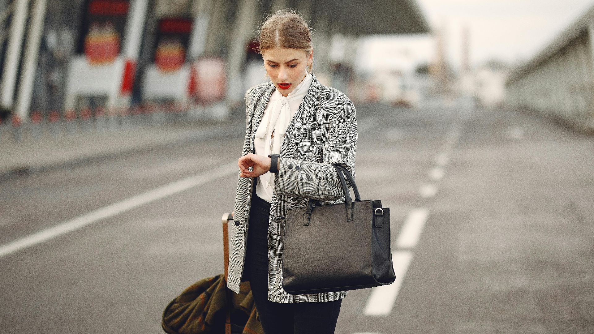 Pensive female passenger in formal outfit checking time on wristwatch while walking along asphalt road to airport terminal with suitcase and handbag during business trip