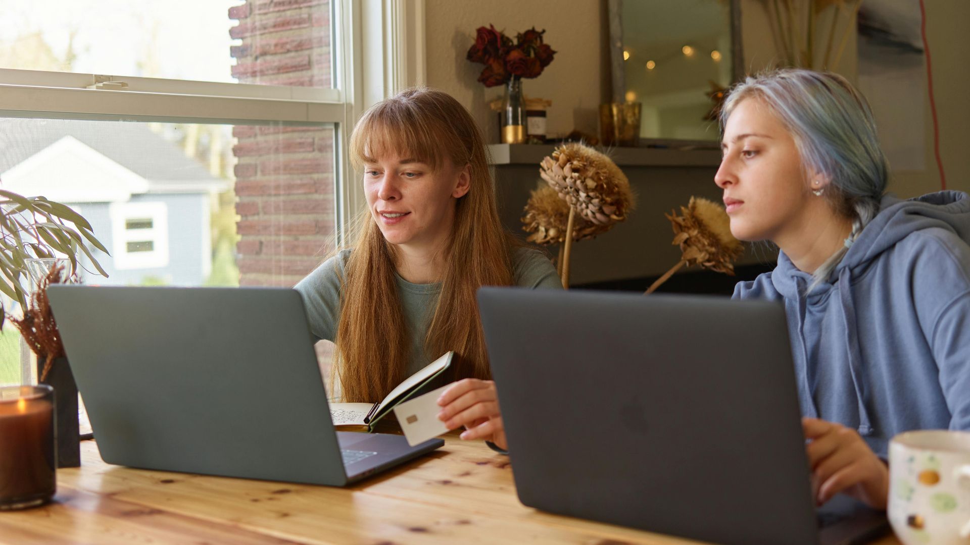Two young women working together at a wooden table using laptops. Cozy indoor setting.