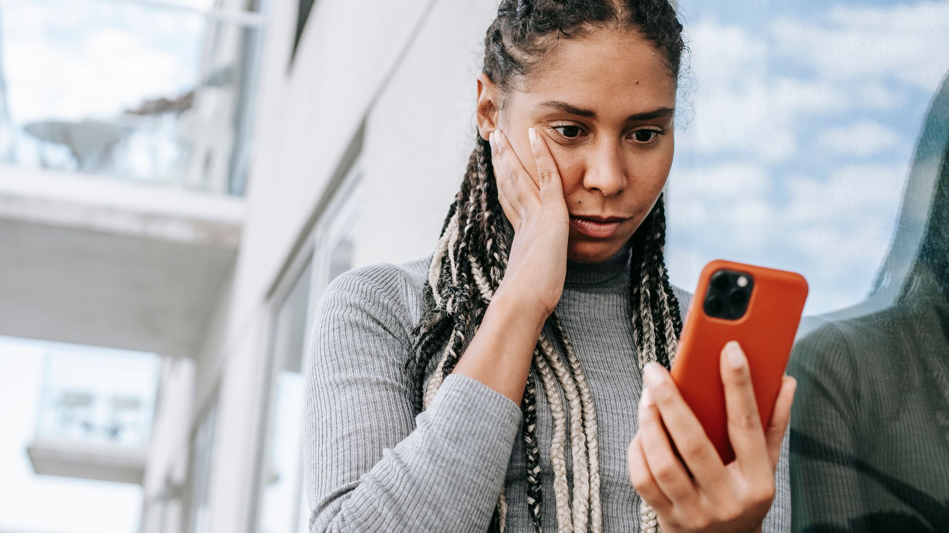 Crop serious African American female with dreadlocks touching cheek in contemplation and browsing mobile phone on street
