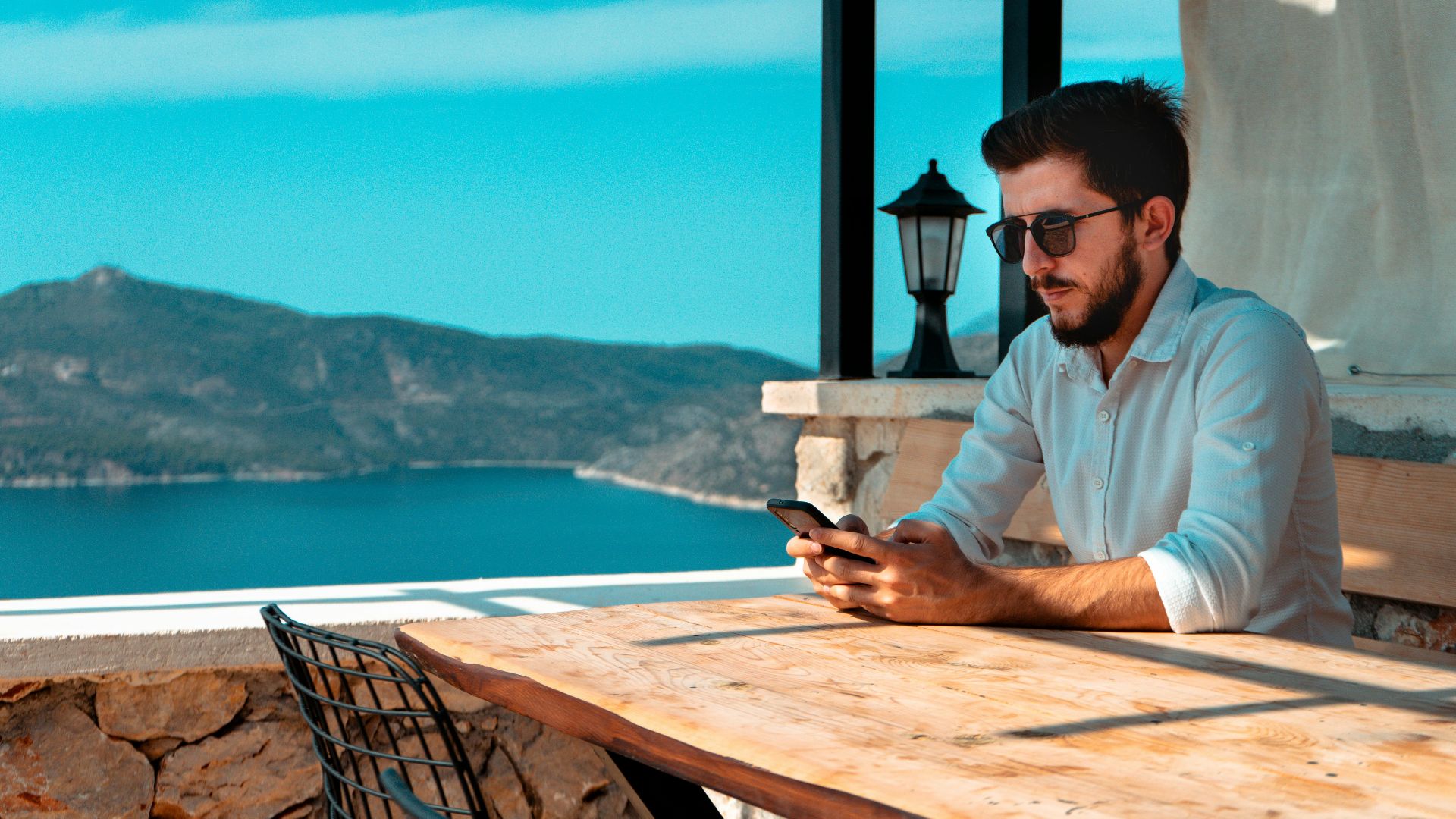 A man using a cellphone while sitting at an outdoor cafe in Antalya, Türkiye, with a mountain view.