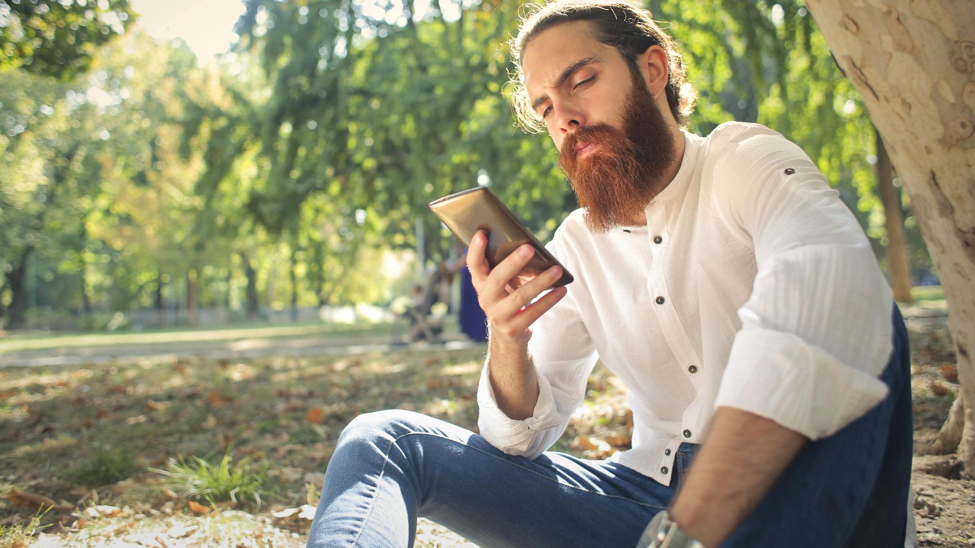 A bearded man in a white shirt uses a smartphone while sitting outdoors in a park.