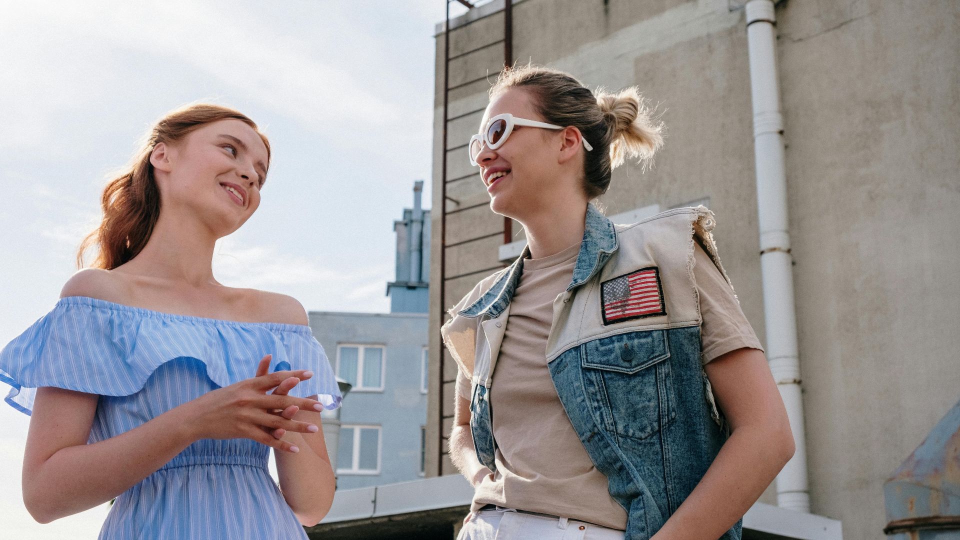 Two women smiling and chatting on a sunlit urban rooftop in summer.