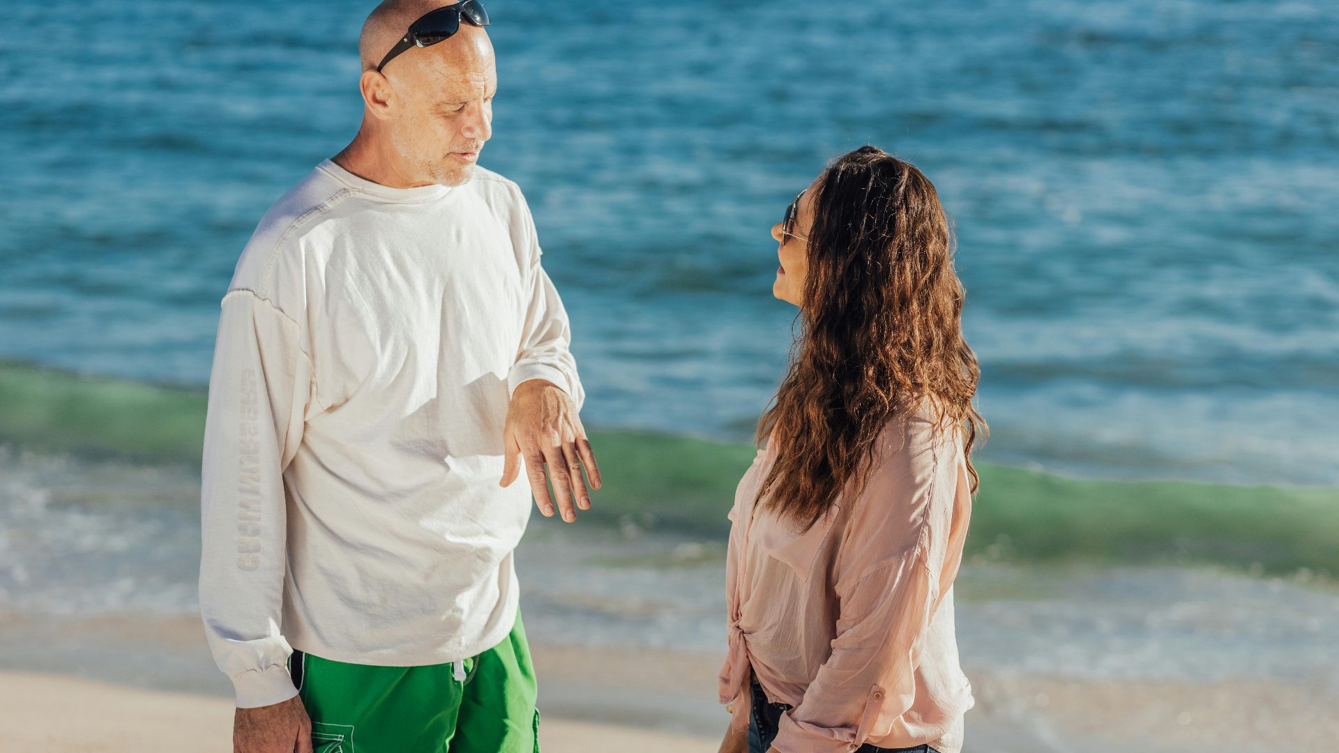 Two adults enjoy a casual conversation on a sunny beach with the ocean as a backdrop.