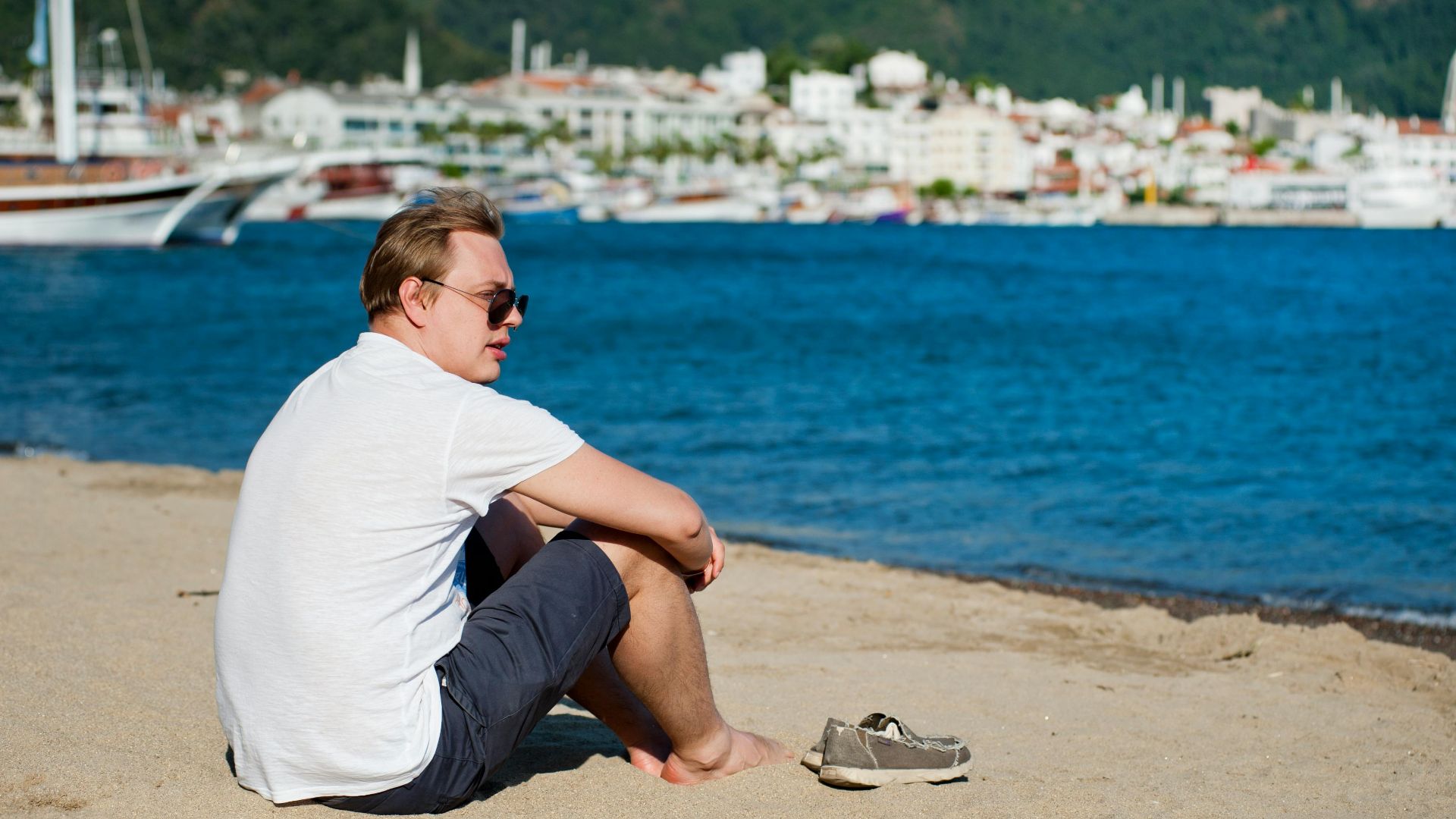 A man sits on a sandy beach, enjoying the sunny day by the ocean with yachts in the background.