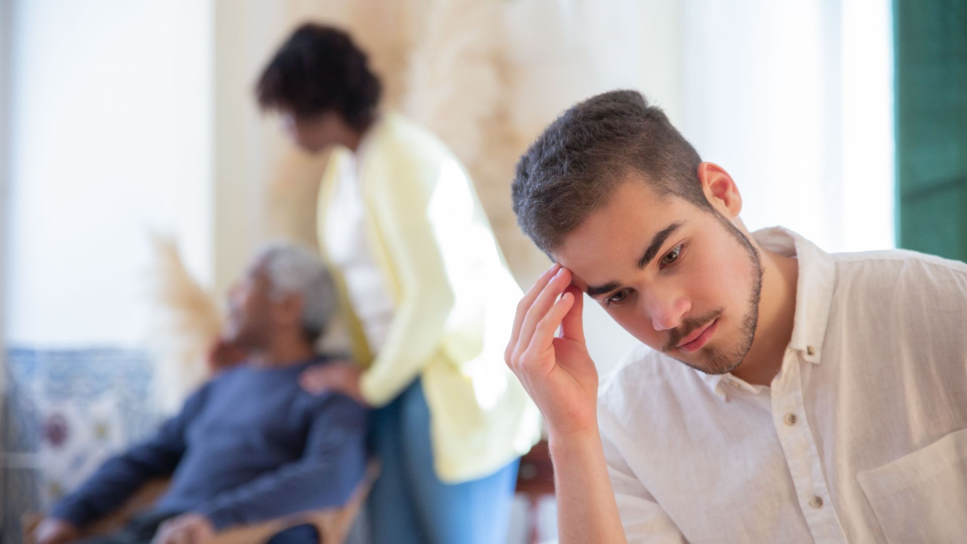 Young man in thoughtful pose indoors with family members in the background, expressing contemplation and care.