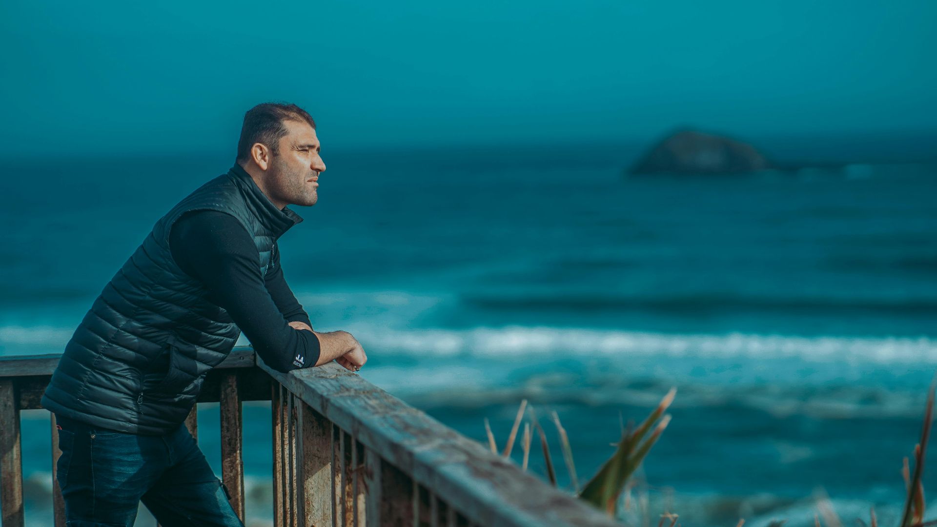 A man leans on a railing, gazing at the ocean with a thoughtful expression.