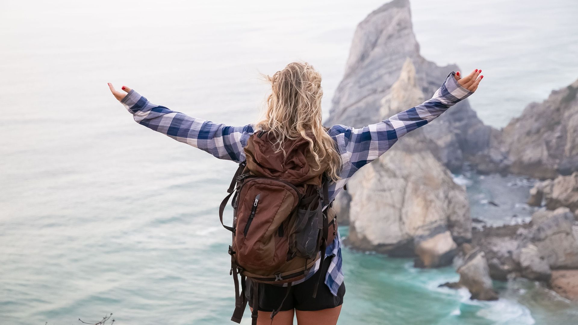 Woman with backpack stands on mountainside with arms wide open, overlooking scenic ocean view.