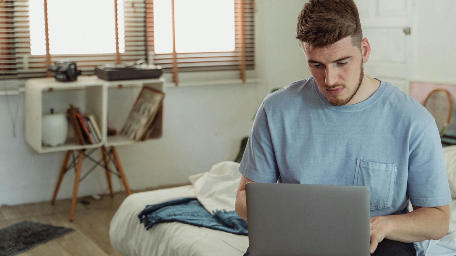 A bearded man in a blue t-shirt focused on a laptop in a sunlit bedroom.