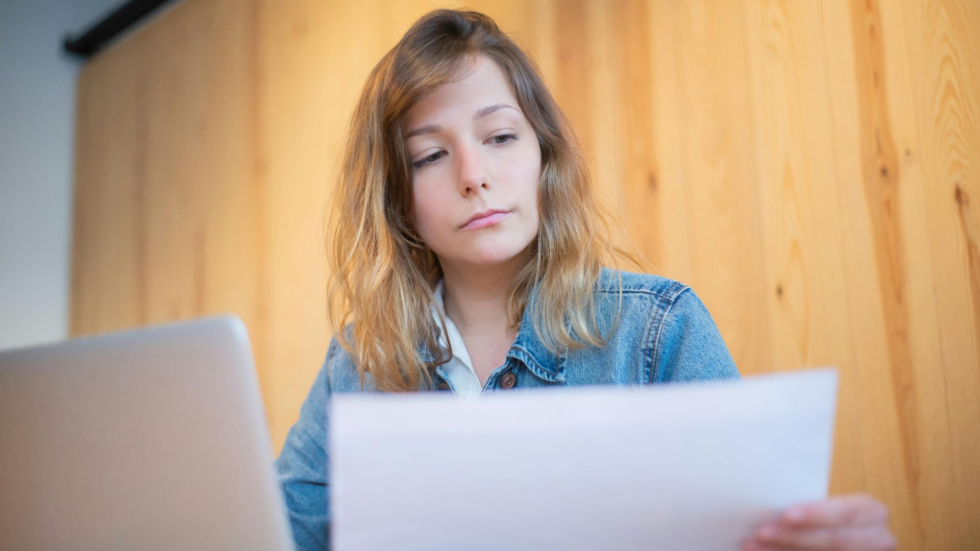 Businesswoman in a denim jacket analyzing documents at a wooden table in a cozy workspace.