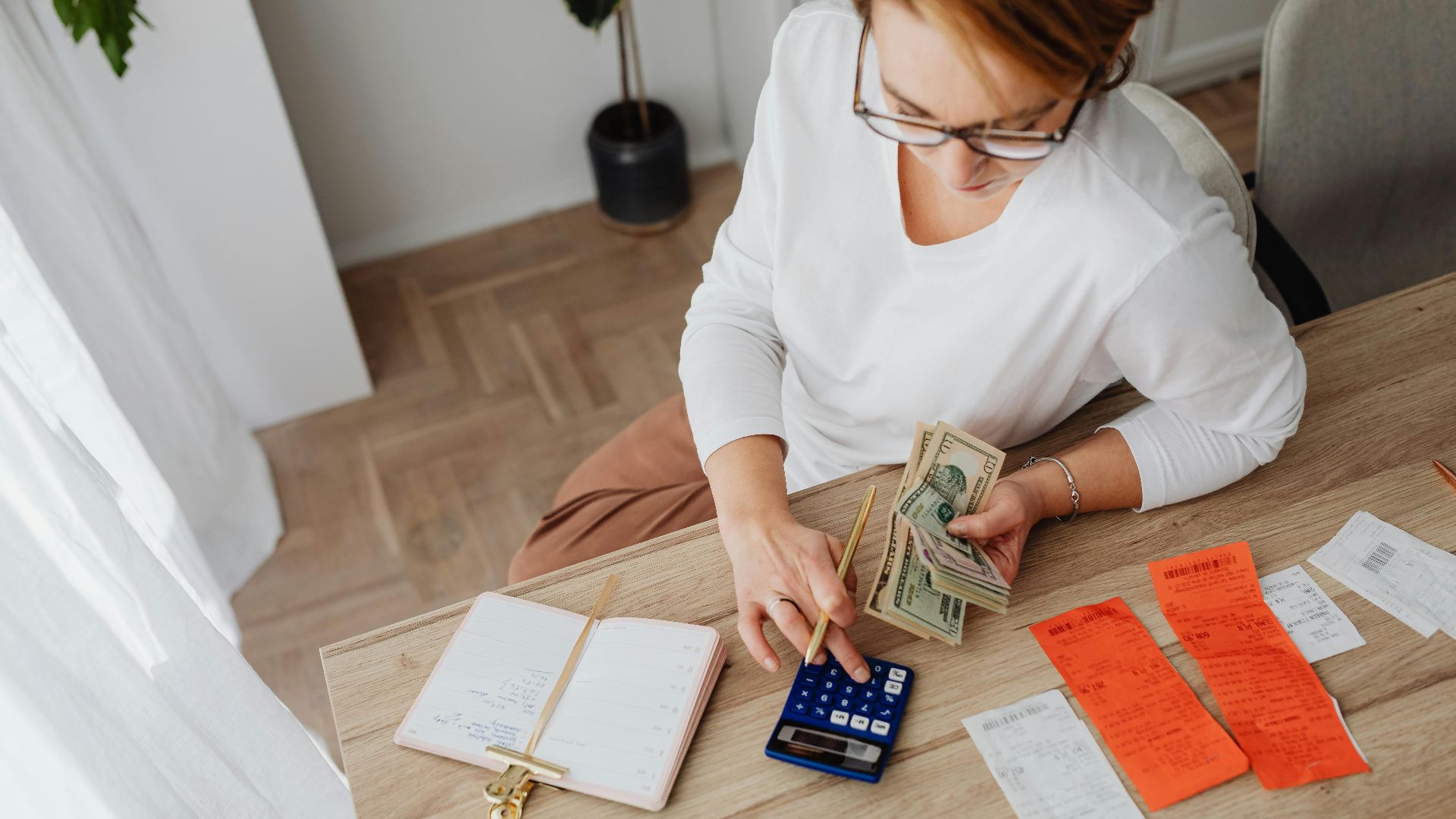Top view of a woman managing finances with a calculator, cash, and bills in a home setting.