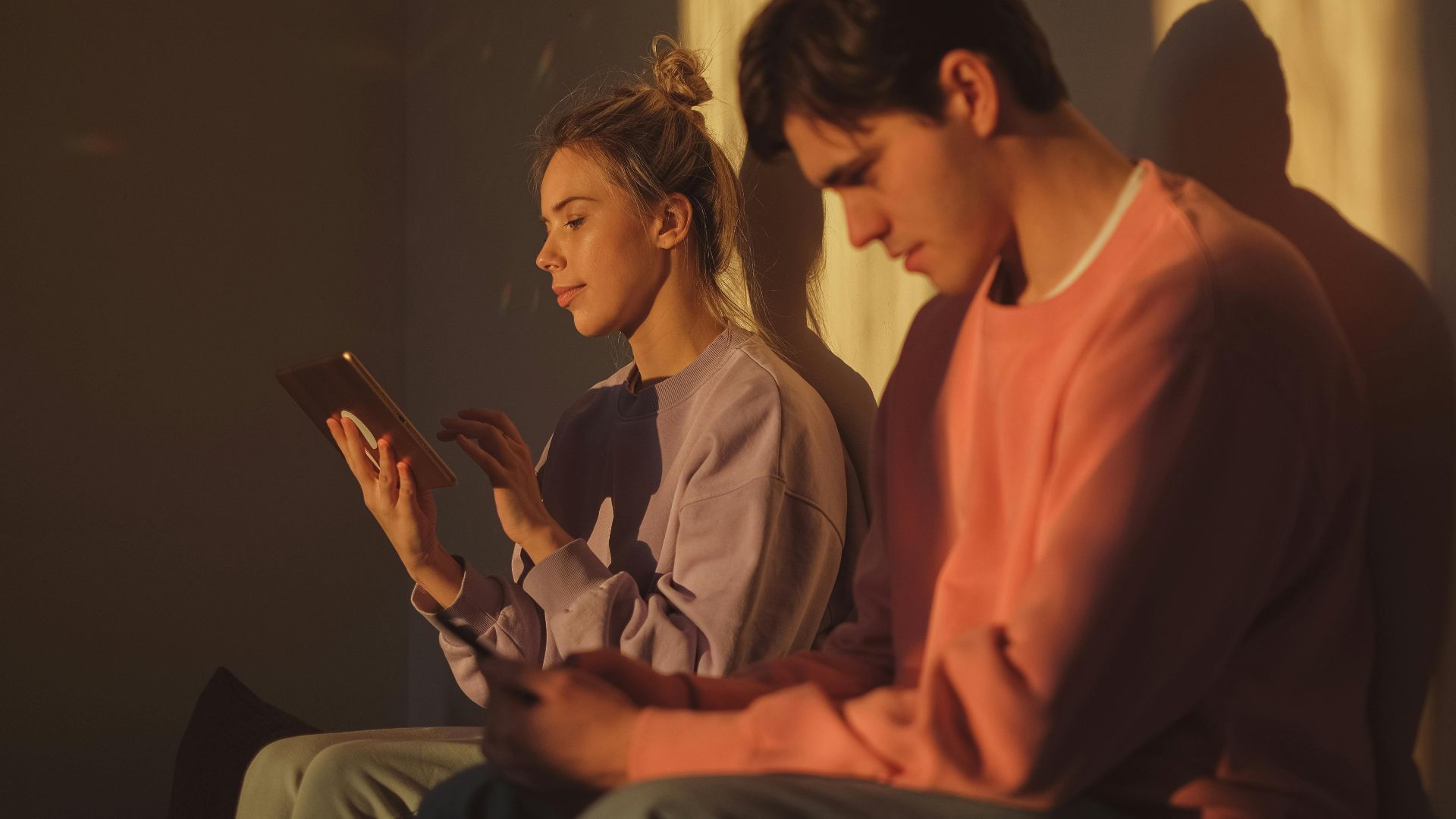 A young couple sitting together indoors, engaged with electronic devices in warm sunlight.