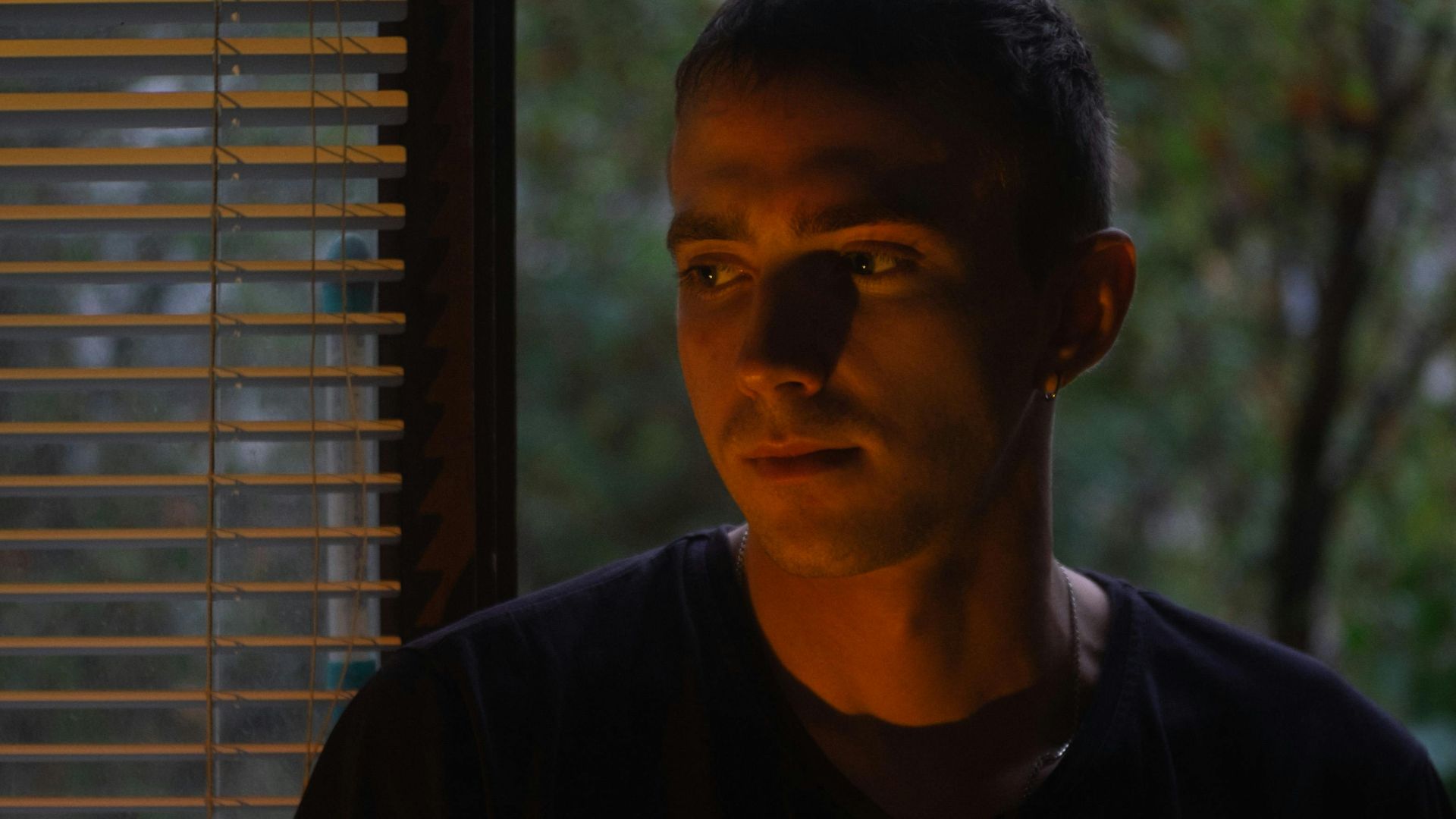 A young man stands by a window, casting dramatic shadows on his face.