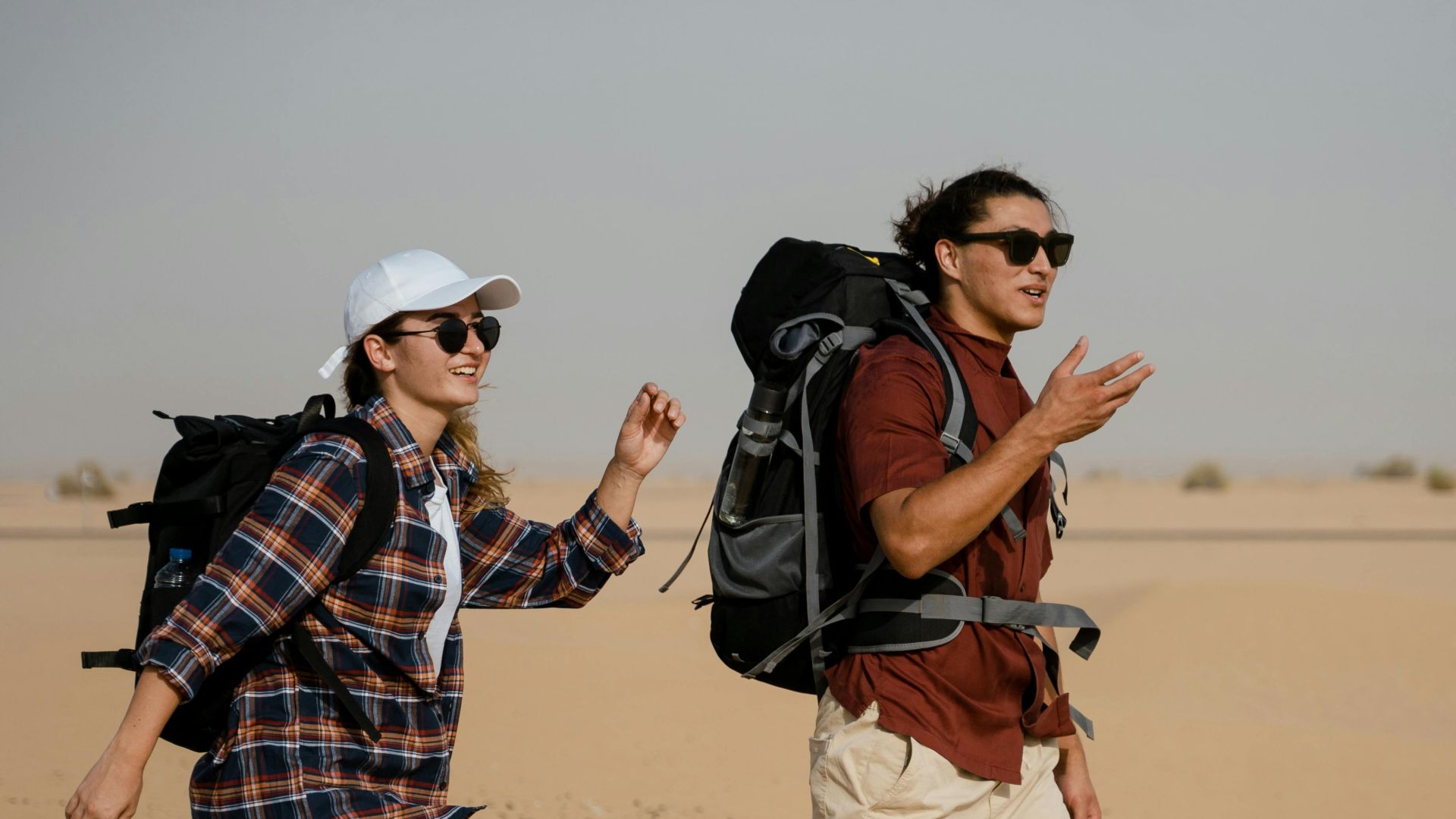 Two young adults hiking with backpacks across an open desert on a sunny day, embracing adventure.