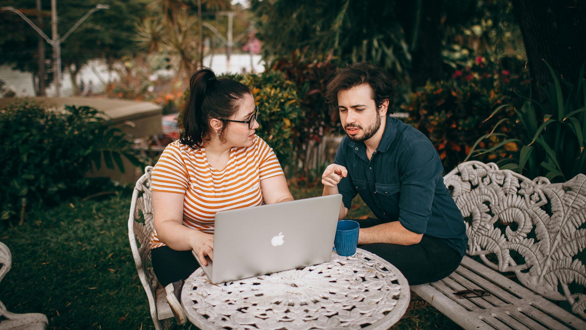 A man and woman working together on a laptop outdoors in a lush garden.