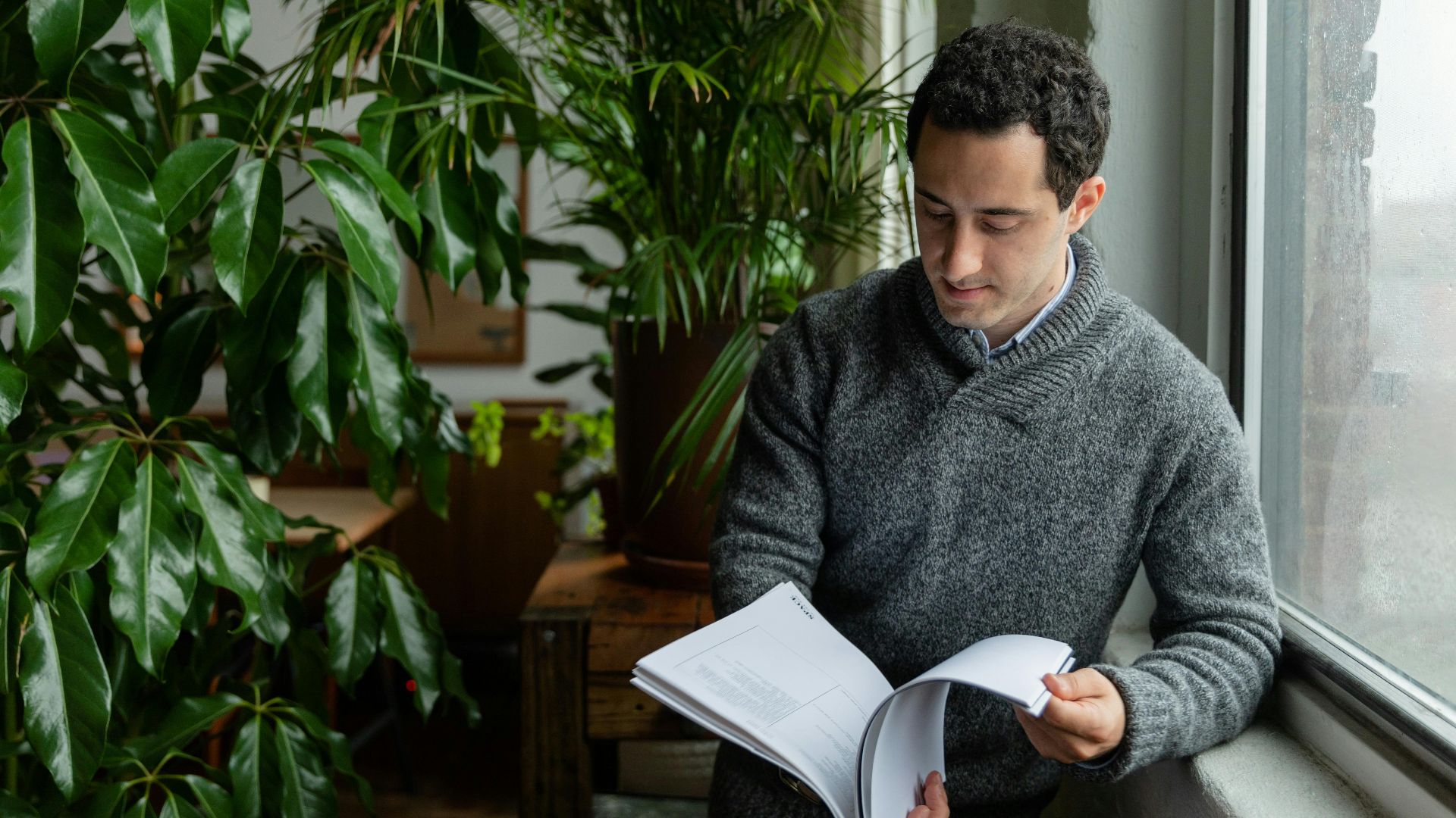 A man in a sweater reads a book next to lush indoor plants by a window.