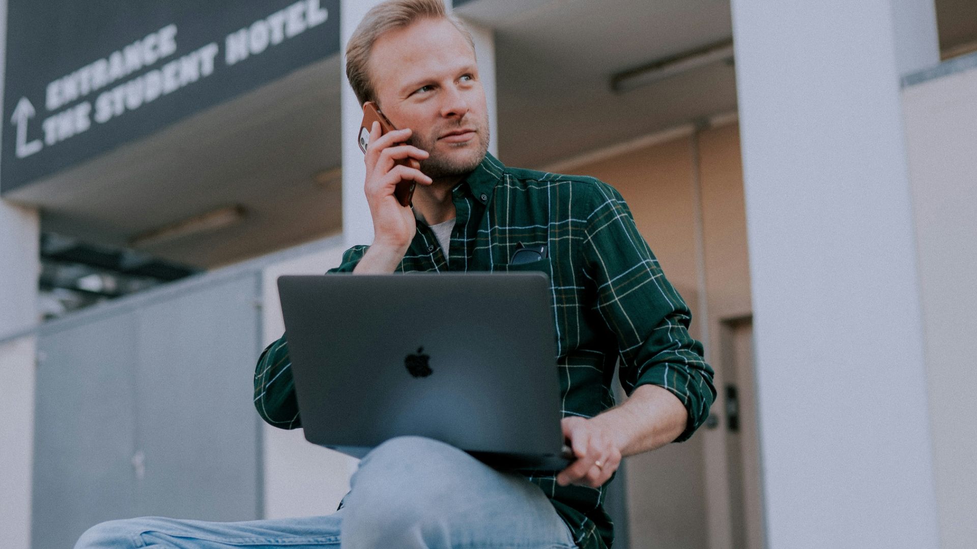 man in green and black plaid dress shirt and blue denim jeans sitting on gray concrete