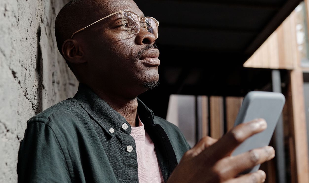 African American man with smartphone pondering against a concrete wall.