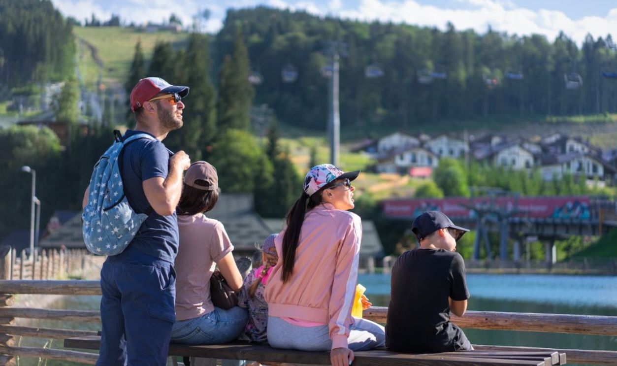 A family enjoys a sunny day sitting on a bench in Bukovel, Ukraine, with scenic mountain views.