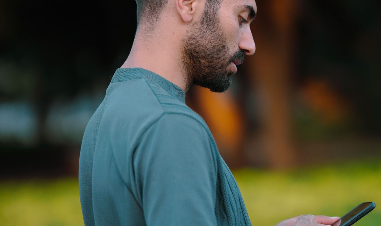 Profile of a man using his smartphone in a lush green park setting during summer.