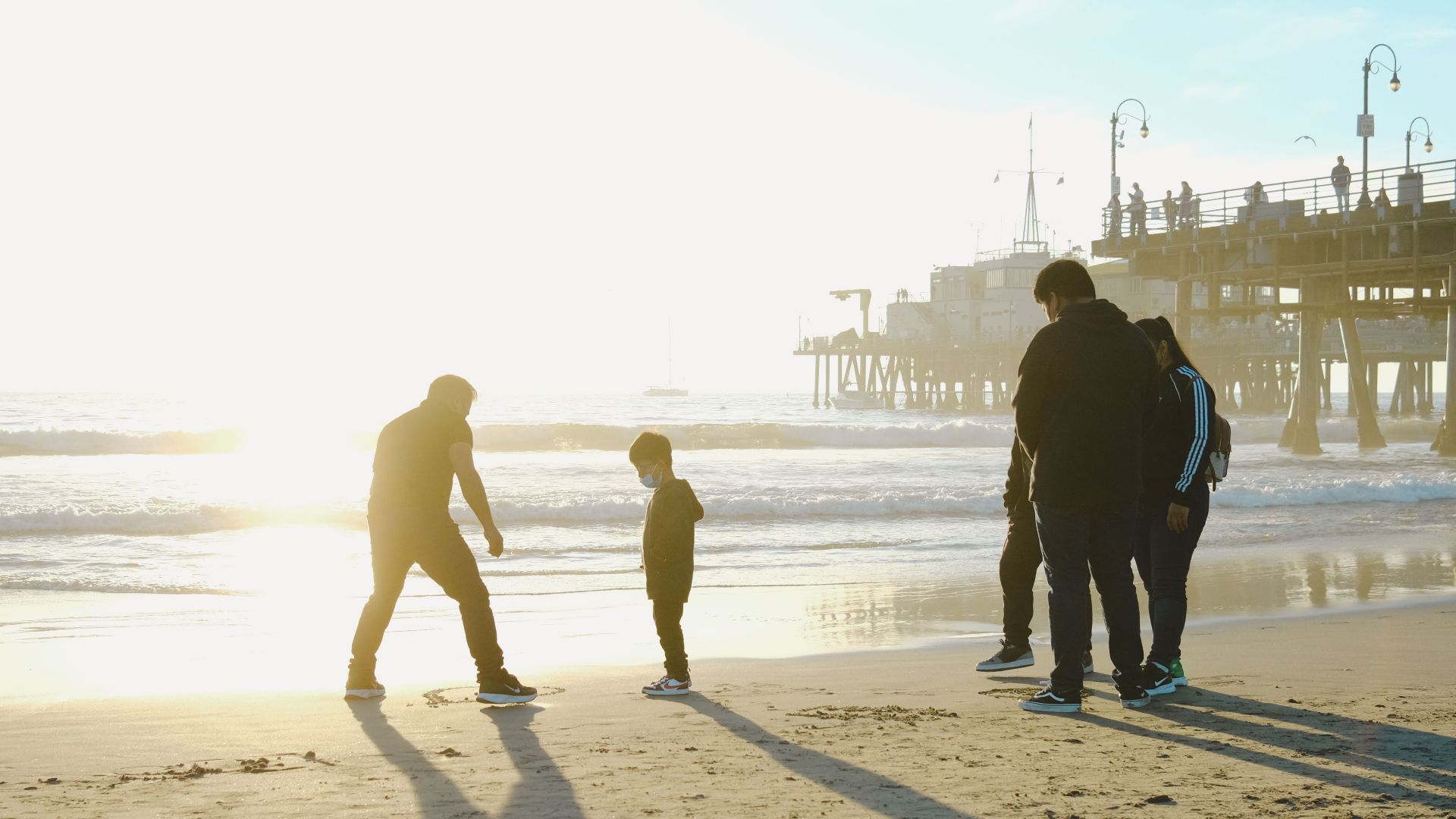 A family enjoys a walk on a beach at sunset, casting long shadows with a pier in the background.