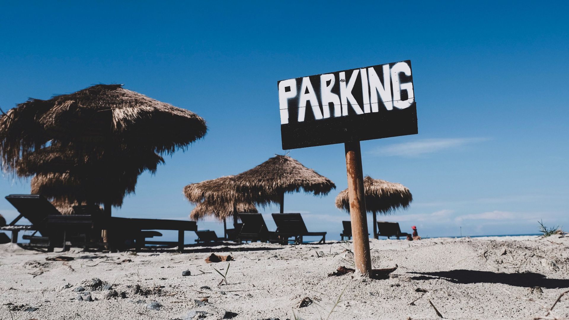 A serene beach scene with a parking sign and straw huts under a clear blue sky.