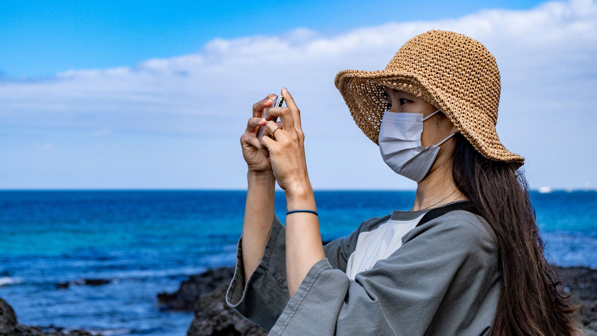A woman wearing a face mask and straw hat takes a picture by the ocean in Jeju, South Korea.