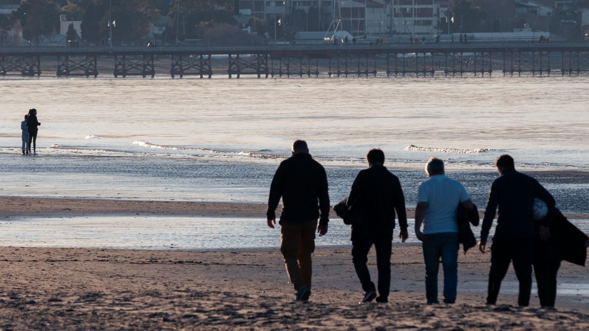 Silhouetted group of men walking along a scenic beach during sunset with cityscape in the background.