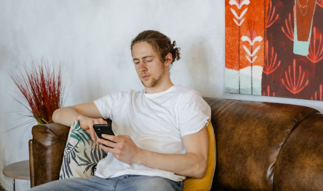Casual young man in white shirt using smartphone on sofa at home