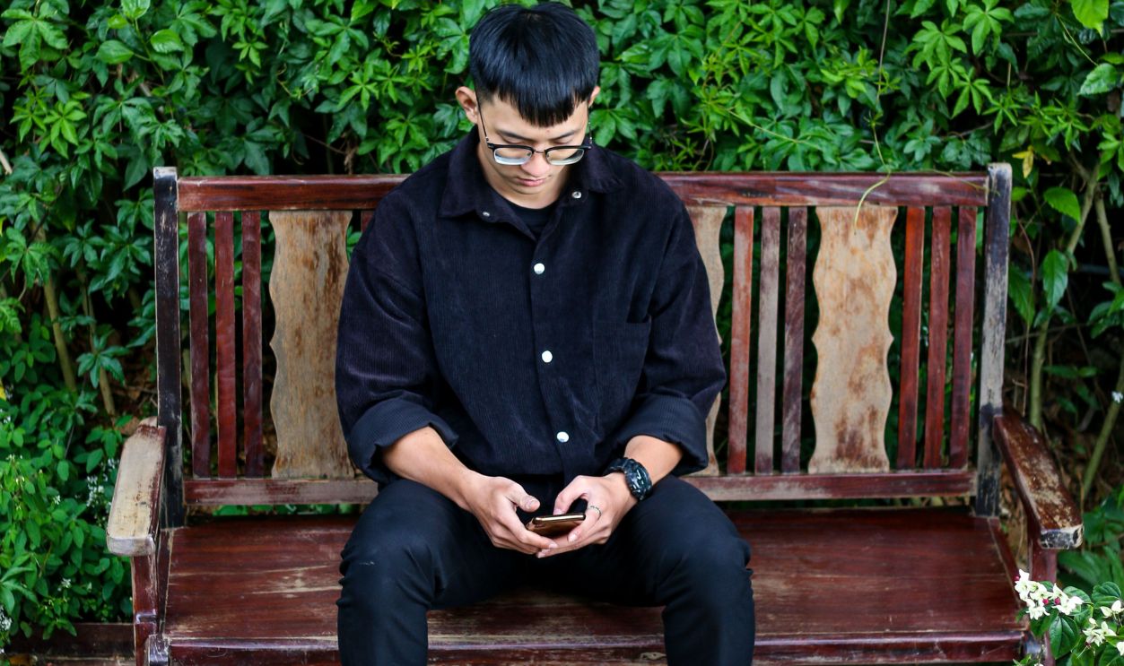 A man sits on a wooden bench in a lush garden, focused on his smartphone.