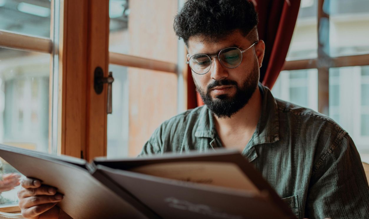Man wearing glasses intently reading a menu at a cozy indoor cafe setting, surrounded by paperwork.
